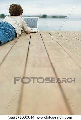 Boy lying on dock, using laptop View Large Photo Image Picture - Boy lying on dock, using laptop. Fotosearch