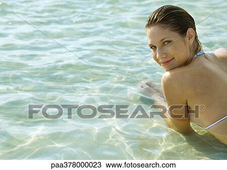 Stock Image - Woman lying in shallow water on beach, looking over shoulder at camera. Fotosearch