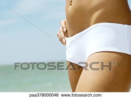 Stock Photo - Woman standing in front of sea, close-up of abdomen. Fotosearch