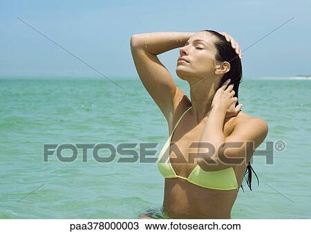 Stock Image - Woman standing in sea, eyes closed and hands in hair. Fotosearch