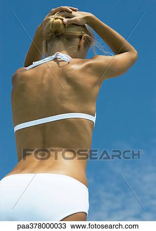Stock Image - Woman standing in sun in bikini, hands behind head, low angle rear view. Fotosearch