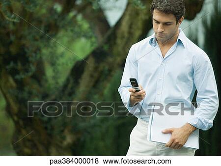 Stock Photo - Man using cell phone outdoors, holding documents. Fotosearch