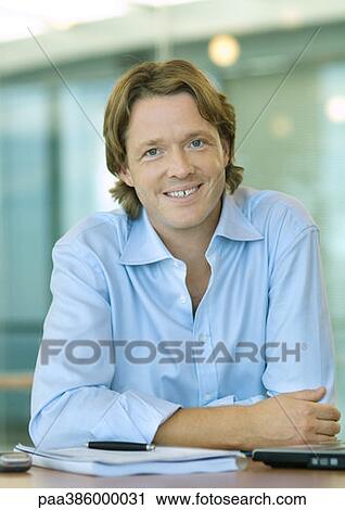 Businessman sitting at desk, smiling at camera, portrait View Large Photo Image Stock Image - Businessman sitting at desk, smiling at camera, portrait. Fotosearch