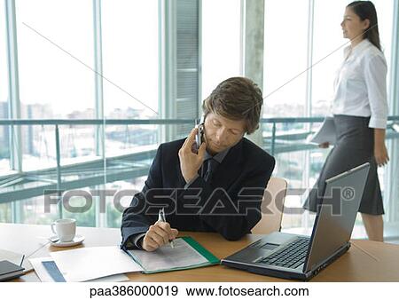 Businessman using cell phone at desk while female office worker walks by in background View Large Photo Image Stock Photo - Businessman using cell phone at desk while female office worker walks by in background. Fotosearch