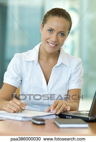 Stock Image - Businesswoman sitting at desk, writing, smiling at camera. Fotosearch