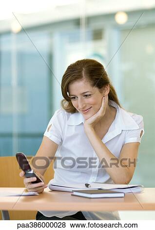 Stock Photo - Woman sitting at desk, smiling and looking at cell phone. Fotosearch