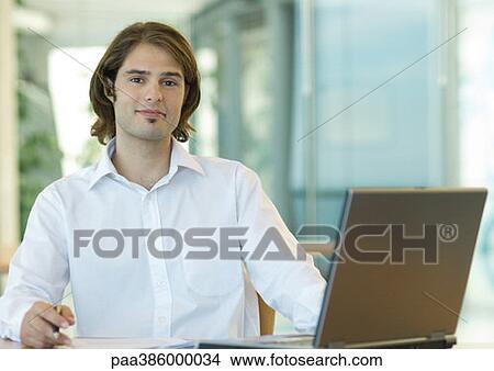 Young businessman sitting at desk, smiling at camera View Large Photo Image Picture - Young businessman sitting at desk, smiling at camera. Fotosearch