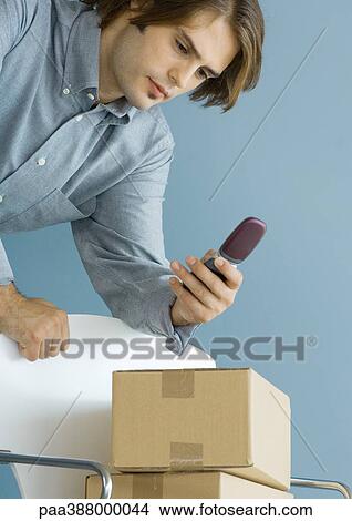 Man leaning against chair, looking at cell phone, cardboard boxes stacked on chair View Large Photo Image Picture - Man leaning against chair, looking at cell phone, cardboard boxes stacked on chair. Fotosearch