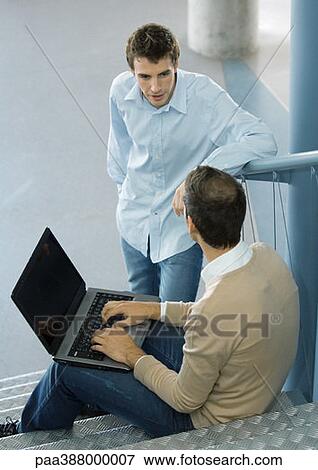 Stock Photo - Man sitting on steps, using laptop, speaking to second man. Fotosearch