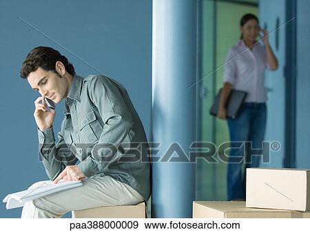 Young man sitting on cardboard box using cell phone, young woman entering in background View Large Photo Image Stock Photo - Young man sitting on cardboard box using cell phone, young woman entering in background. Fotosearch