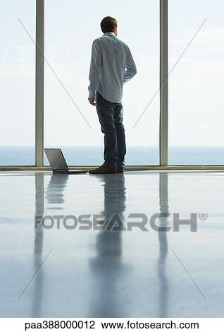 Young man standing by window, laptop on floor, full length View Large Photo Image Stock Image - Young man standing by window, laptop on floor, full length. Fotosearch