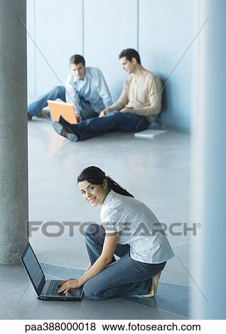 Young woman crouching sitting on floor, using laptop, men sitting in background View Large Photo Image Stock Photo - Young woman crouching sitting on floor, using laptop, men sitting in background. Fotosearch
