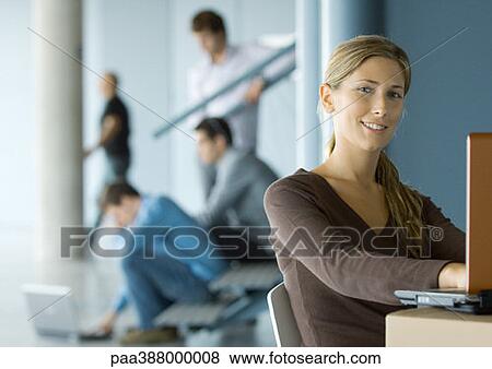 Young woman using laptop, smiling at camera, busy office in background View Large Photo Image Stock Photo - Young woman using laptop, smiling at camera, busy office in background. Fotosearch