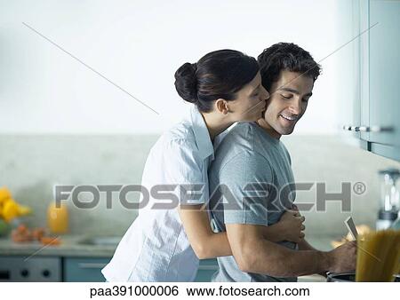 Woman kissing man from behind in the kitchen View Large Photo Image Stock Photograph - Woman kissing man from behind in the kitchen. Fotosearch