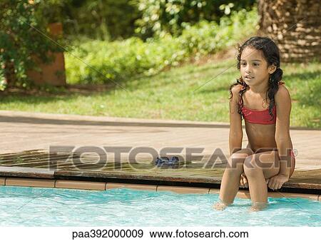 Stock Photo - Girl sitting on edge of swimming pool with feet in water. Fotosearch