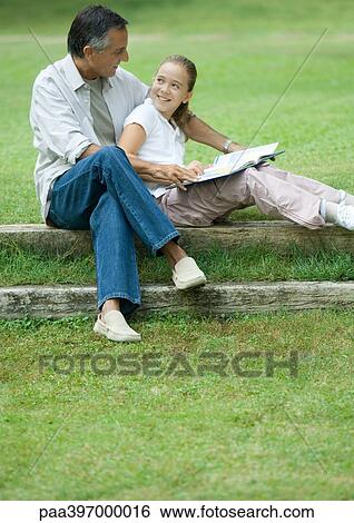 Stock Photograph - Mature man reading with granddaughter outdoors. Fotosearch