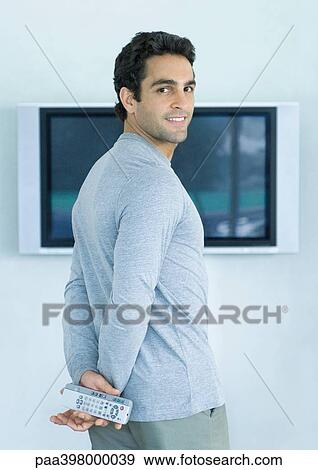 Man standing in front of widescreen TV, holding two remote controls, smiling over shoulder at camera View Large Photo Image Stock Photo - Man standing in front of widescreen TV, holding two remote controls, smiling over shoulder at camera. Fotosearch