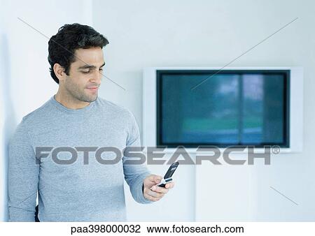 Man standing, looking at cell phone, widescreen TV on wall in background View Large Photo Image Stock Image - Man standing, looking at cell phone, widescreen TV on wall in background. Fotosearch