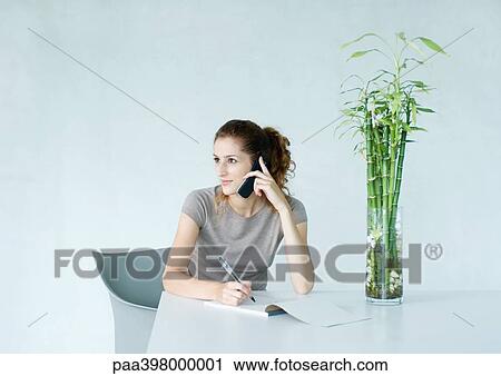 Stock Image - Woman sitting at table with phone to ear, taking notes . Fotosearch