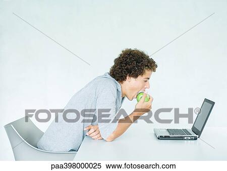 Young man sitting at table, eating apple and using laptop View Large Photo Image Stock Photography - Young man sitting at table, eating apple and using laptop. Fotosearch