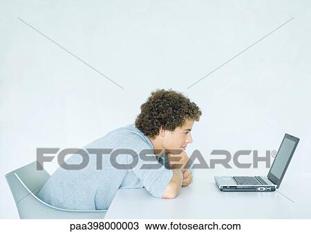 Young man sitting, leaning on desk, facing laptop computer View Large Photo Image Stock Image - Young man sitting, leaning on desk, facing laptop computer. Fotosearch