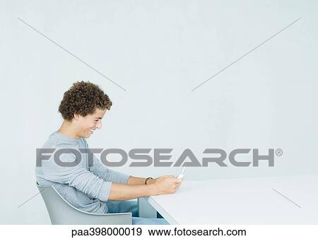 Stock Photo - Young man sitting, looking at cell phone. Fotosearch