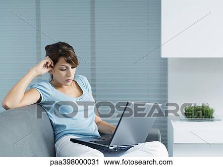 Young woman sitting on sofa, using laptop View Large Photo Image Stock Image - Young woman sitting on sofa, using laptop. Fotosearch