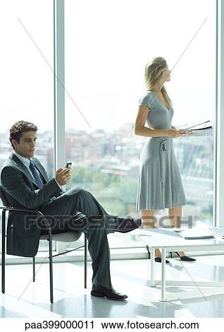 Businessman and woman waiting in lobby, man checking cell phone, woman standing in background, holding magazine View Large Photo Image Stock Image - Businessman and woman waiting in lobby, man checking cell phone, woman standing in background, holding magazine. Fotosearch