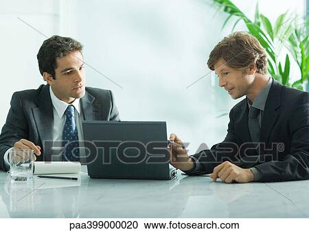 Two businesmen sitting at table, looking at laptop View Large Photo Image Stock Image - Two businesmen sitting at table, looking at laptop. Fotosearch