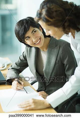 Woman sitting as desk, pointing to document and smiling at second woman View Large Photo Image Stock Image - Woman sitting as desk, pointing to document and smiling at second woman. Fotosearch