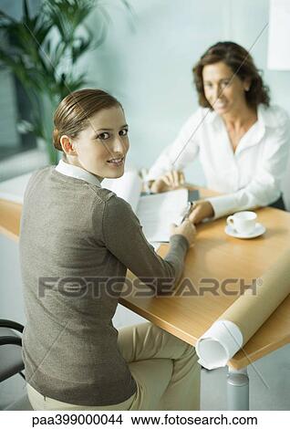 Woman sitting at table across from architect, smiling over shoulder at camera View Large Photo Image Picture - Woman sitting at table across from architect, smiling over shoulder at camera. Fotosearch