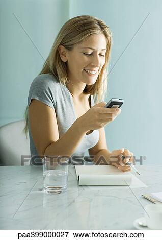 Woman sitting at table, looking at cell phone, smiling View Large Photo Image Stock Photo - Woman sitting at table, looking at cell phone, smiling. Fotosearch