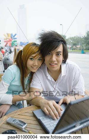 Picture - Teenage couple sitting outdoors, using laptop, smiling at camera. Fotosearch
