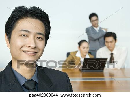 Businessman smiling at camera, team using laptop in background View Large Photo Image Picture - Businessman smiling at camera, team using laptop in background. Fotosearch