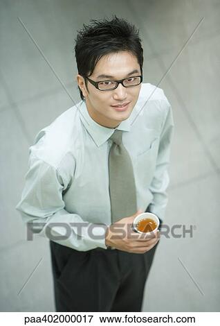 Businessman standing, holding cup of tea, smiling at camera, high angle view View Large Photo Image Stock Photo - Businessman standing, holding cup of tea, smiling at camera, high angle view. Fotosearch