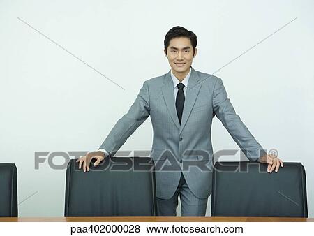 Businessman standing with hands on backs of chairs, smiling at camera, three quarter length View Large Photo Image Stock Photo - Businessman standing with hands on backs of chairs, smiling at camera, three quarter length. Fotosearch
