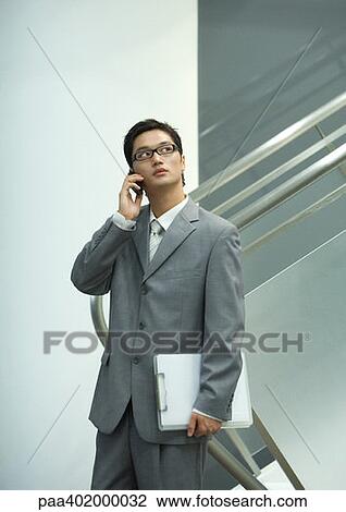 Stock Image - Businessman using cell, holding file, looking up. Fotosearch