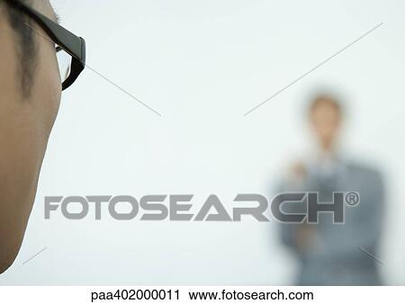 Stock Image - Man wearing glasses, side of face, facing business man in blurred background. Fotosearch