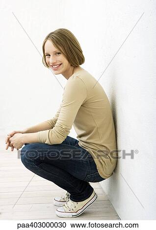 Stock Image - Teenage girl squatting, leaning against wall, portrait. Fotosearch