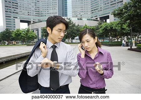 Stock Photo - Two young business executives in office park, woman using cell phone and man holding up arm with watch. Fotosearch