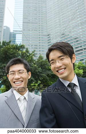 Stock Image - Two young businessmen, laughing, office building in background. Fotosearch