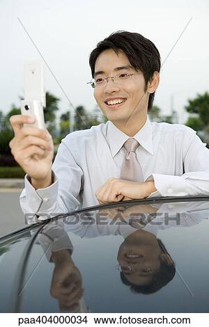 Young businessman leaning against car, holding up cell phone, smiling View Large Photo Image Picture - Young businessman leaning against car, holding up cell phone, smiling. Fotosearch