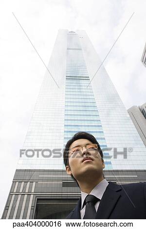 Young businessman standing in front of skyscraper, low angle view View Large Photo Image Stock Photograph - Young businessman standing in front of skyscraper, low angle view. Fotosearch