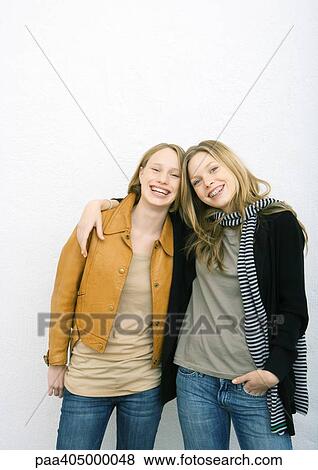 Stock Photo - Two young female friends, one with arm around the other's shoulders, both smiling at camera. Fotosearch
