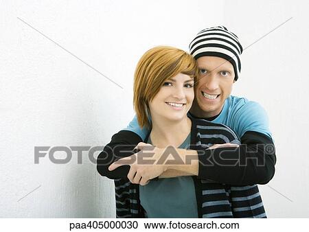Stock Image - Young couple, man standing behind woman with arms around her, both smiling at camera. Fotosearch