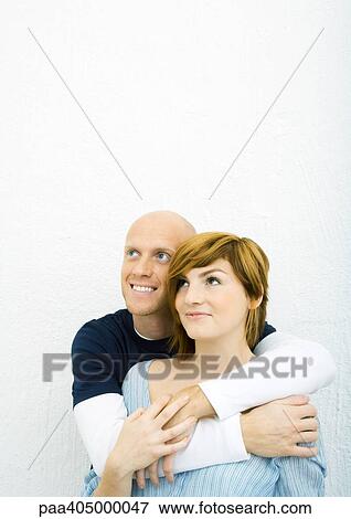 Stock Photo - Young couple, man standing behind woman with arms around her, both looking up. Fotosearch