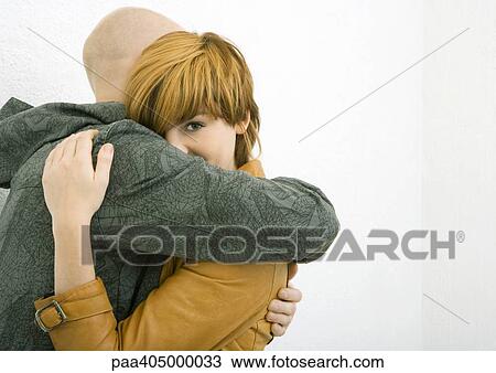 Stock Image - Young man and woman hugging, woman looking over man's arm, looking at camera. Fotosearch