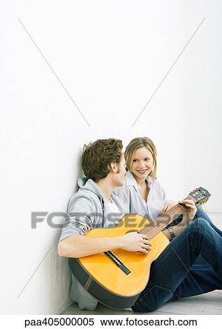 Stock Photography - Young man and woman sitting on ground, man playing guitar. Fotosearch