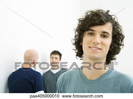 Stock Image - Young man smiling at camera, two young men talking in background. Fotosearch