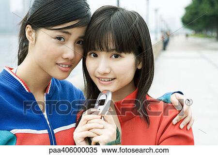 Two female friends, one holding cell phone, smiling at camera View Large Photo Image Stock Photography - Two female friends, one holding cell phone, smiling at camera. Fotosearch